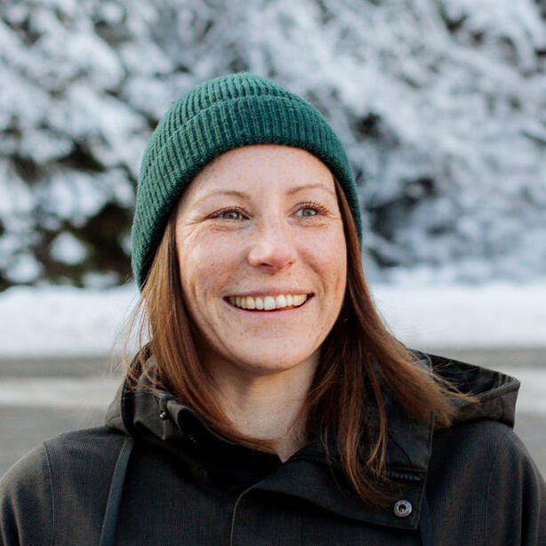 Close-up portrait of Jen, smiling and standing in front of snow-covered trees. She's wearing a dark green beanie and a black winter jacket.