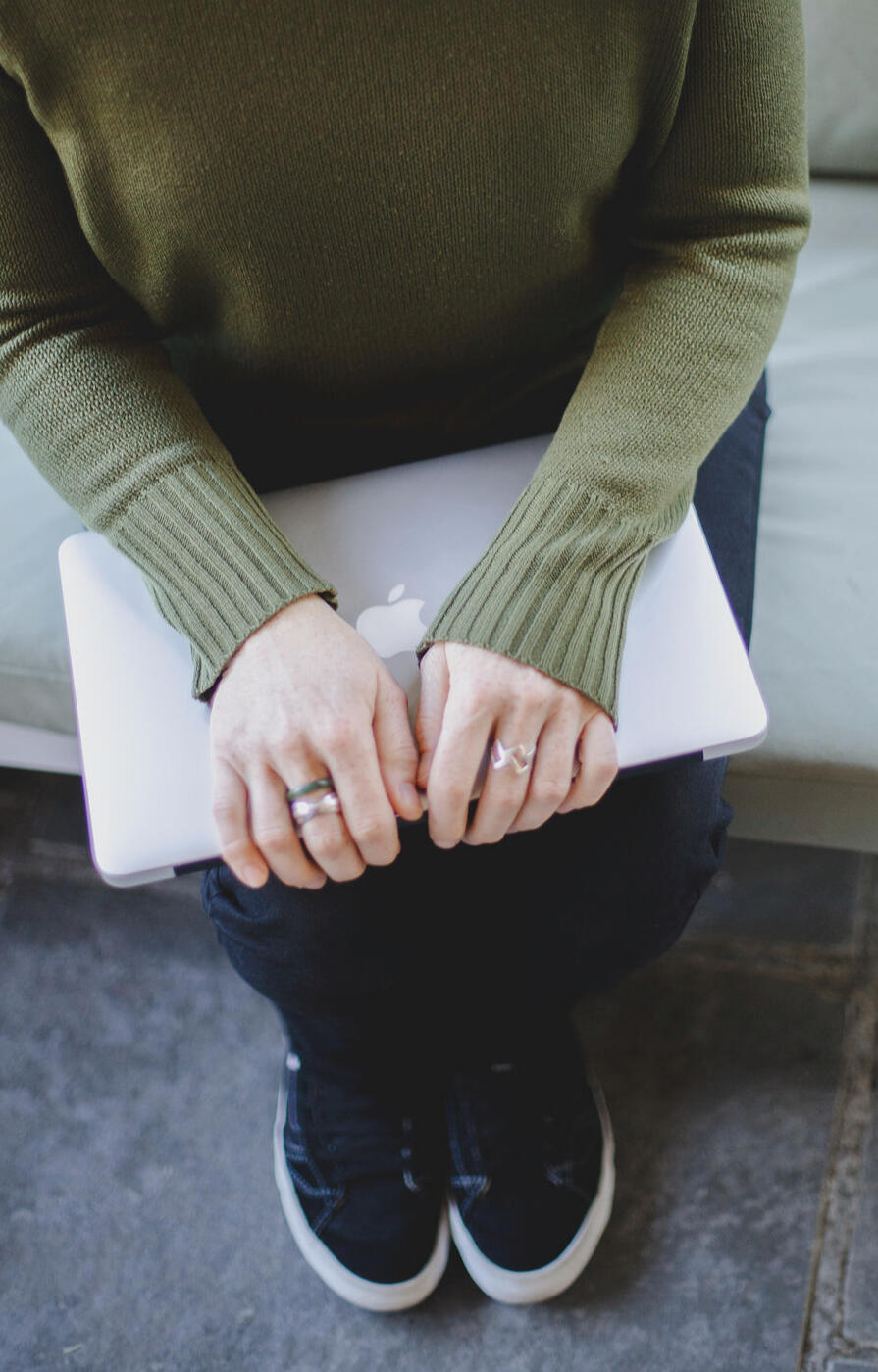 Top-down view of Jen wearing an olive-green knit sweater, dark jeans, and black sneakers. She's holding a closed laptop on her lap with both hands resting on it. Her face is out of frame.