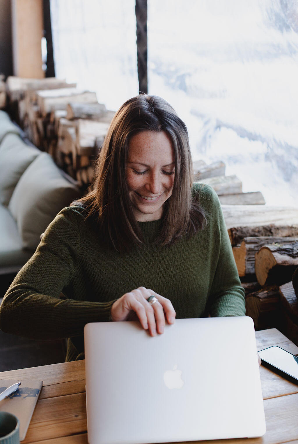 Jen smiling while opening her laptop, sitting at a wooden table, with stacked logs and a snowy window view behind her.