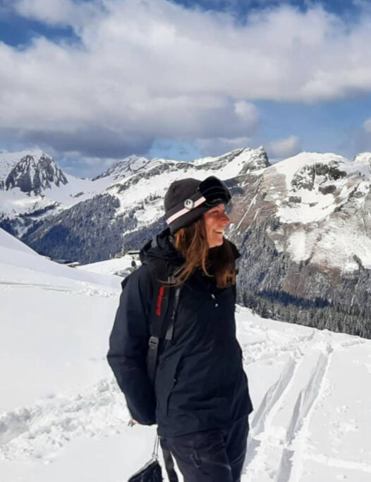 Side-profile image of Jen next to splitboard tracks in the snow in Avoriaz. She's smiling, wearing black clothing, a grey beanie, snowboard goggles with a pink strap, and a backpack. Snow-covered mountains and a partly cloudy sky are in the background.